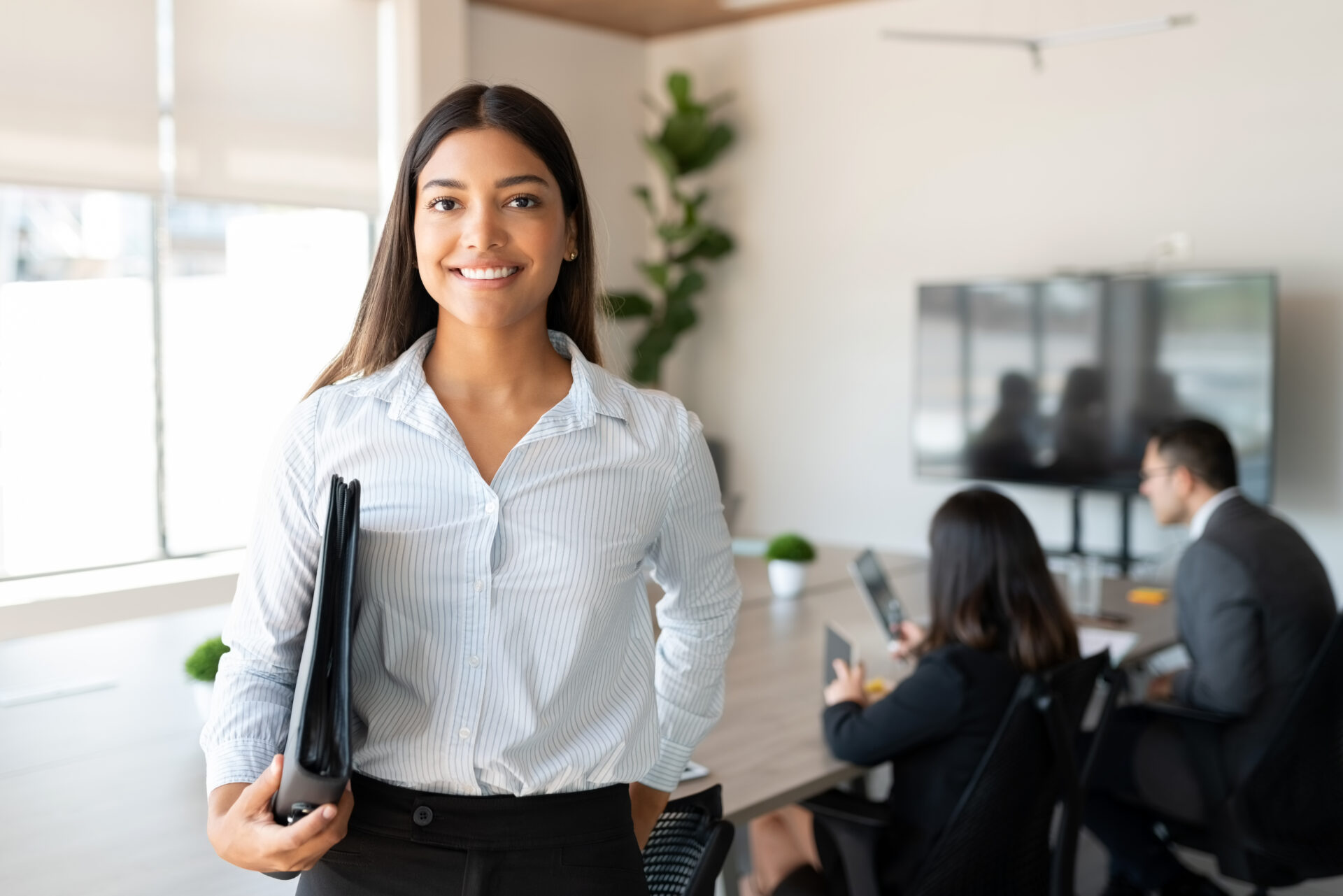 Portrait of hispanic businesswoman with a file standing in meeting room with colleagues disucssing in background